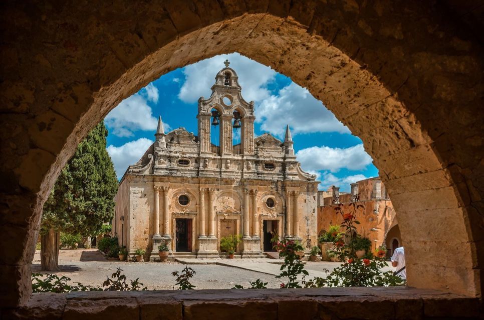 An ornate stone church with a bell tower, viewed from within a dark stone archway on a sunny day.