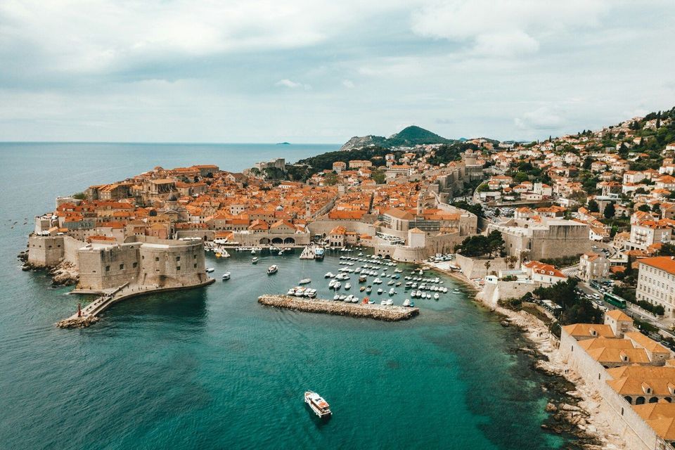 Vista aérea de una ciudad histórica amurallada con tejados de tejas naranjas en una península, con barcos en un pequeño puerto.