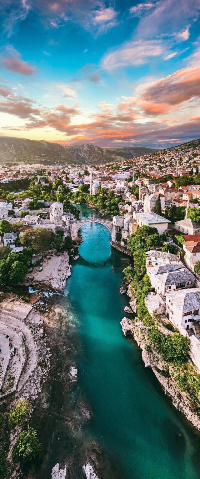 Un puente de piedra arqueado cruza un río turquesa que fluye a través de una ciudad histórica al atardecer, con montañas a lo lejos.
