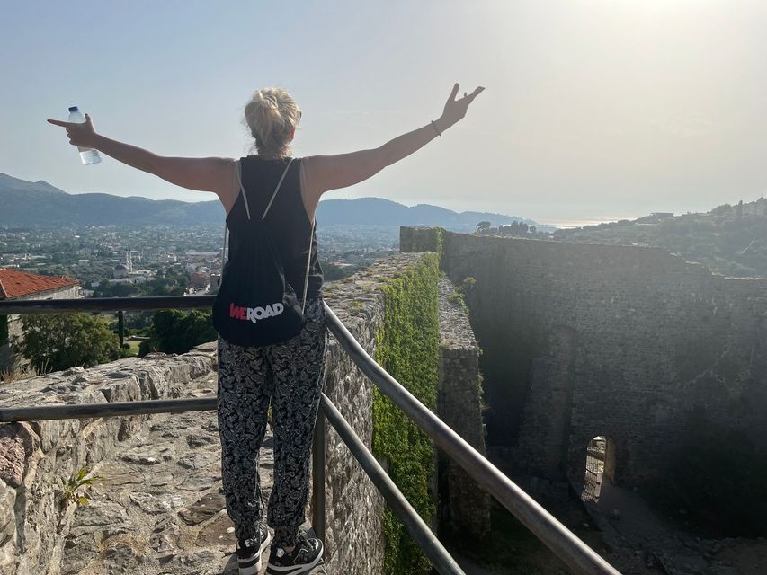 Una mujer con una mochila WeRoad se para en un muro de piedra con los brazos extendidos, contemplando una ciudad costera desde un punto alto.