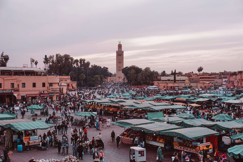 A bustling market square filled with people and green-topped stalls, overlooked by a tall minaret under a pinkish dusk sky.