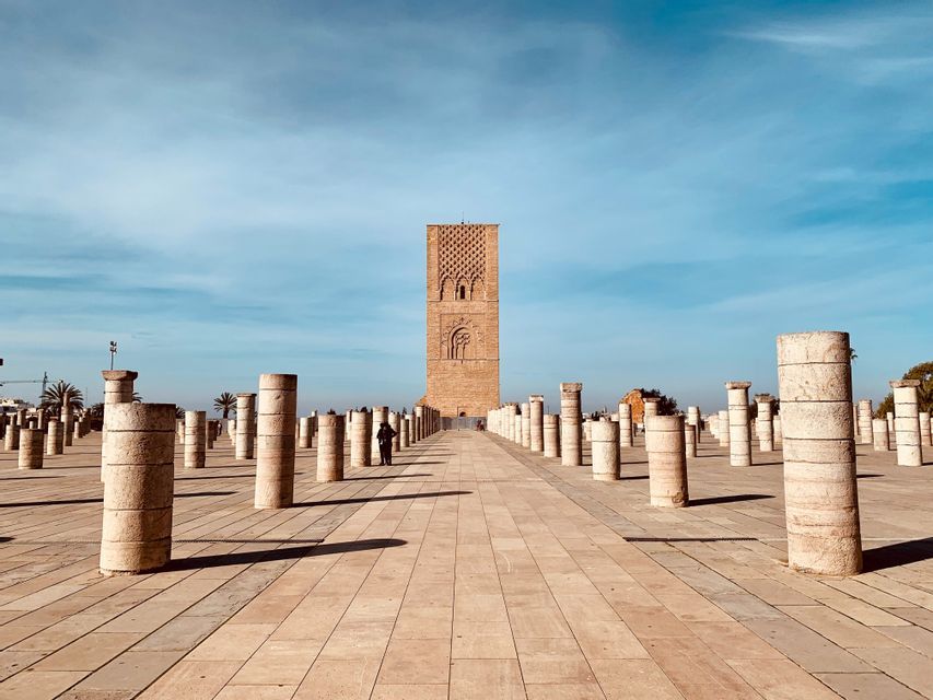 Rows of stone columns fill a wide, paved plaza, leading toward a tall, historic tower under a blue sky.