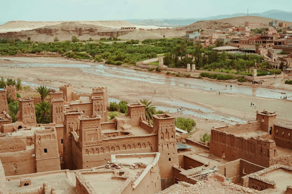 Una vista elevata di una kasbah di mattoni di fango con torri che si affaccia su un ampio letto di fiume, un'oasi verde e distanti colline aride.