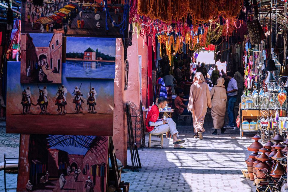 A sun-drenched market alley with people strolling past stalls selling paintings, ceramics, and colorful hanging textiles.