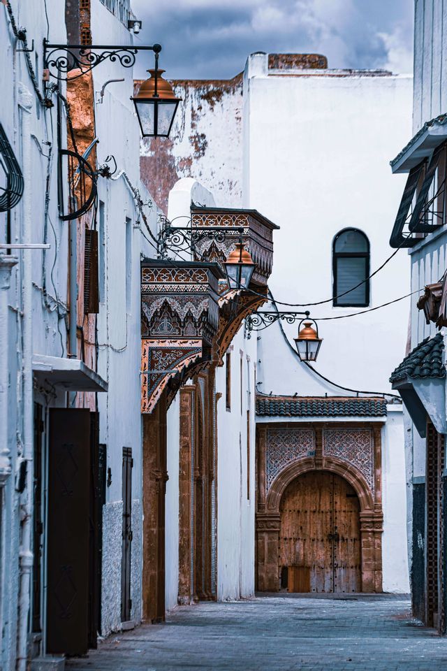 A narrow street with whitewashed buildings, ornate lanterns, and detailed tilework leading to a large arched wooden door.