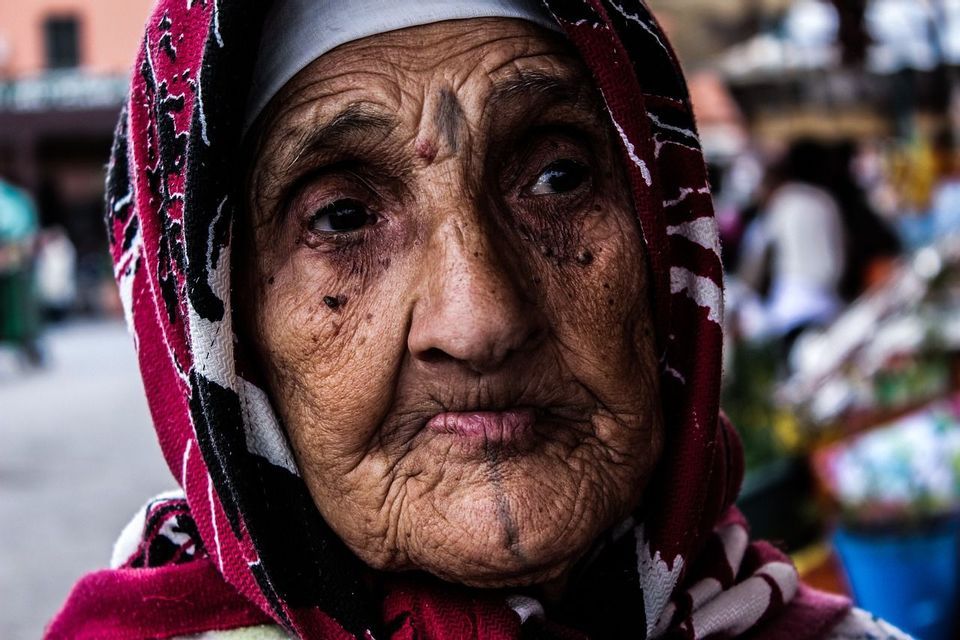 Un ritratto ravvicinato di una donna anziana con la pelle rugosa, che indossa un foulard a motivi rossi, neri e bianchi.