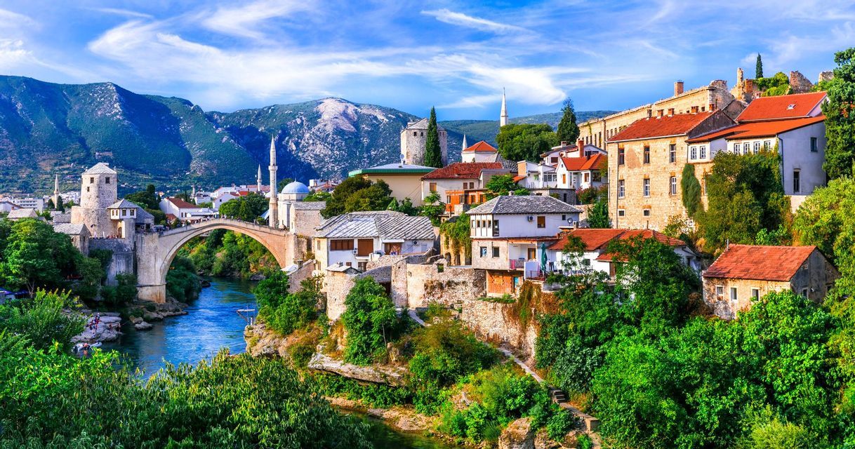 A coastal town with red-tiled roofs and a stone bell tower on the edge of a turquoise bay, with mountains in the distance.