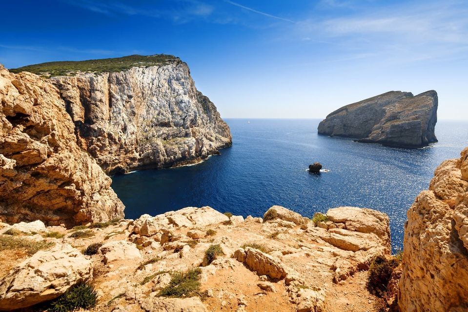 Vista da una scogliera rocciosa che si affaccia su un mare blu profondo, con grandi formazioni rocciose e un cielo azzurro limpido.