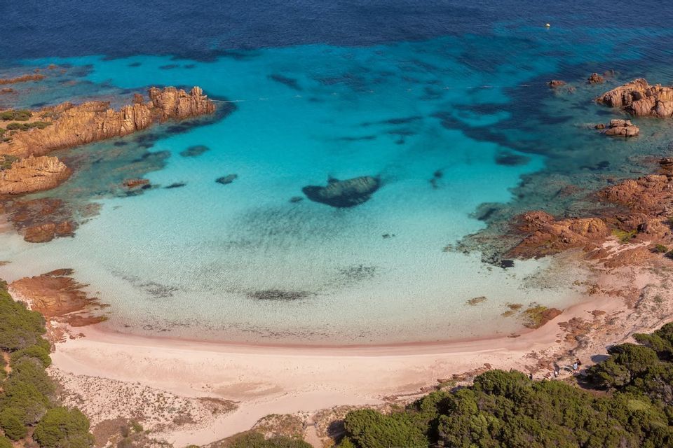 Una vista aerea di una caletta appartata con una spiaggia sabbiosa, incorniciata da rocce rosse e acqua cristallina color turchese.