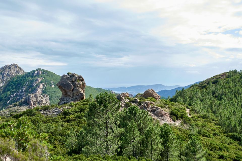 Una grande formazione rocciosa si erge da un fianco montuoso verde e boscoso, affacciandosi su colline lontane e sul mare sotto un cielo nuvoloso.