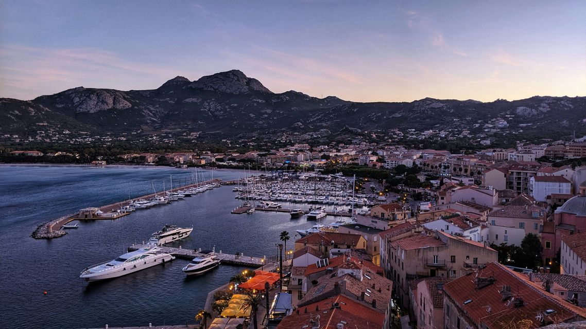Una vista sopraelevata di un porto pieno di barche e yacht, con una città costiera e montagne sullo sfondo al tramonto.