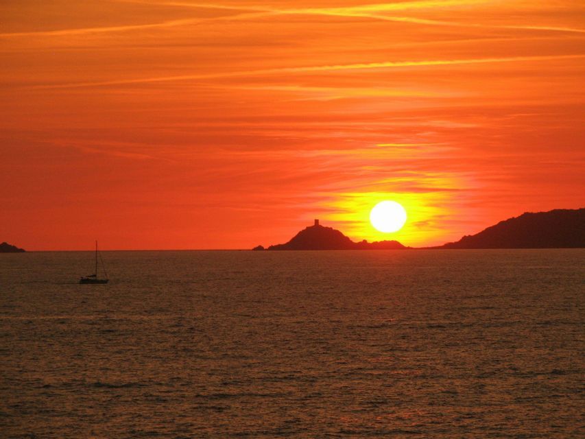 Il sole tramonta sull'oceano, stagliando un'isola con una torre e una barca a vela sull'acqua, tutto sotto un cielo arancione.