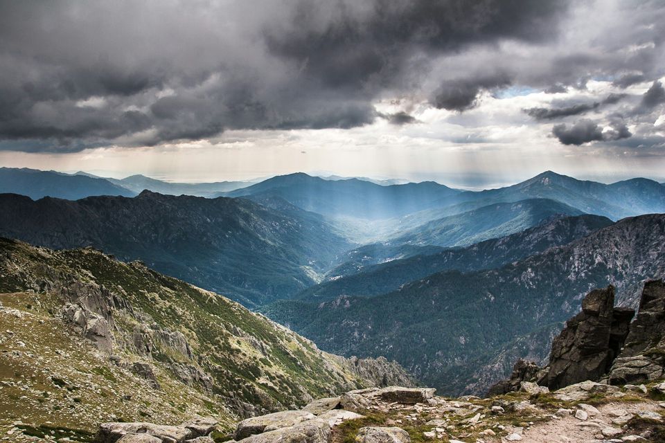 Raggi di sole squarciano nuvole scure, illuminando una valle lussureggiante tra strati di montagne lontane.