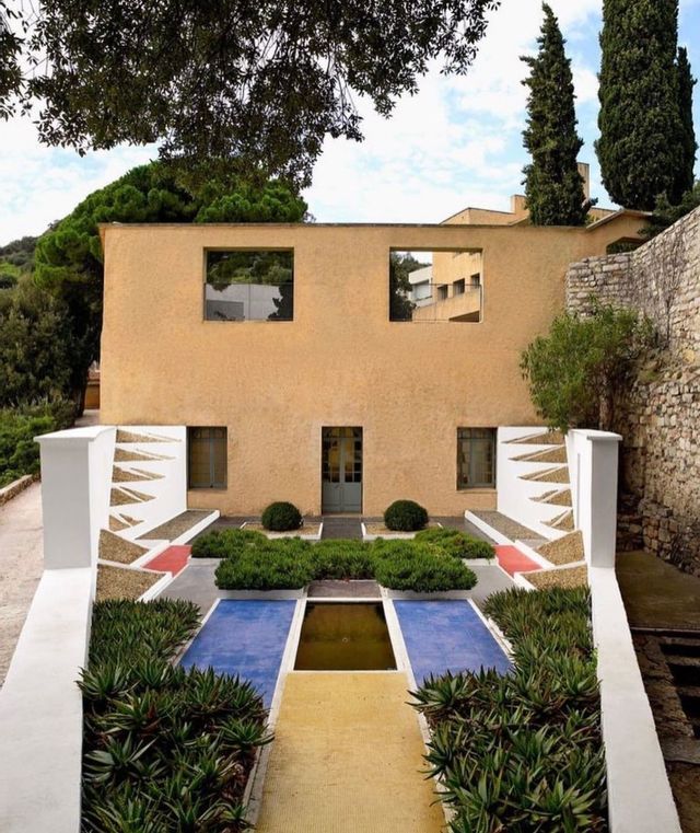 Un edificio modernista con un giardino simmetrico e geometrico caratterizzato da piastrelle colorate, arbusti verdi e una fontana centrale.
