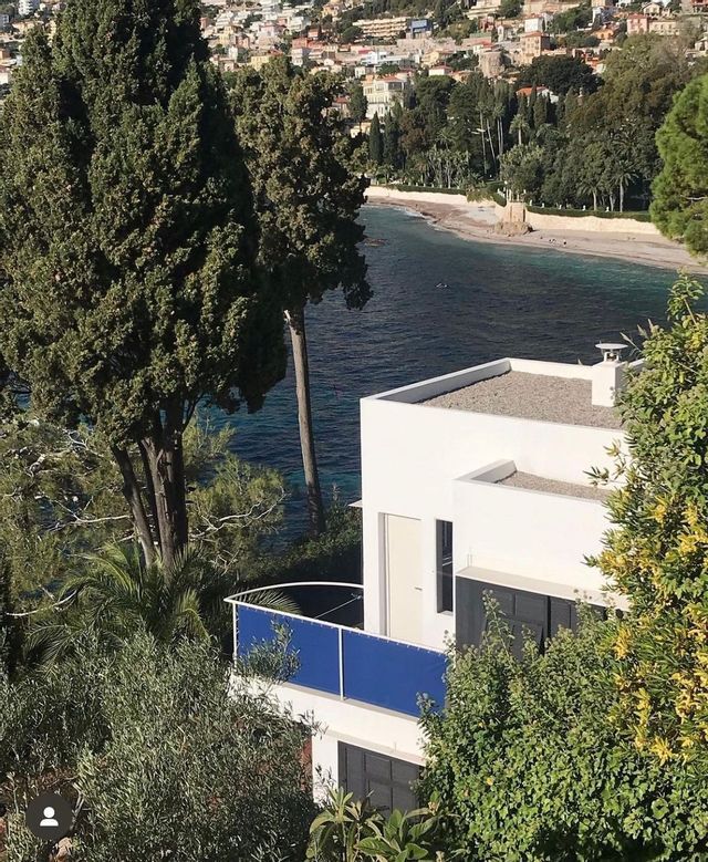 Una casa bianca moderna con un balcone blu, incorniciata da alberi, si affaccia su una baia con una spiaggia sabbiosa e una città costiera