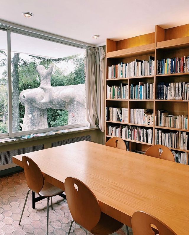 Una sala biblioteca con un lungo tavolo in legno e librerie, caratterizzata da un'ampia finestra che si affaccia su una scultura astratta in pietra.