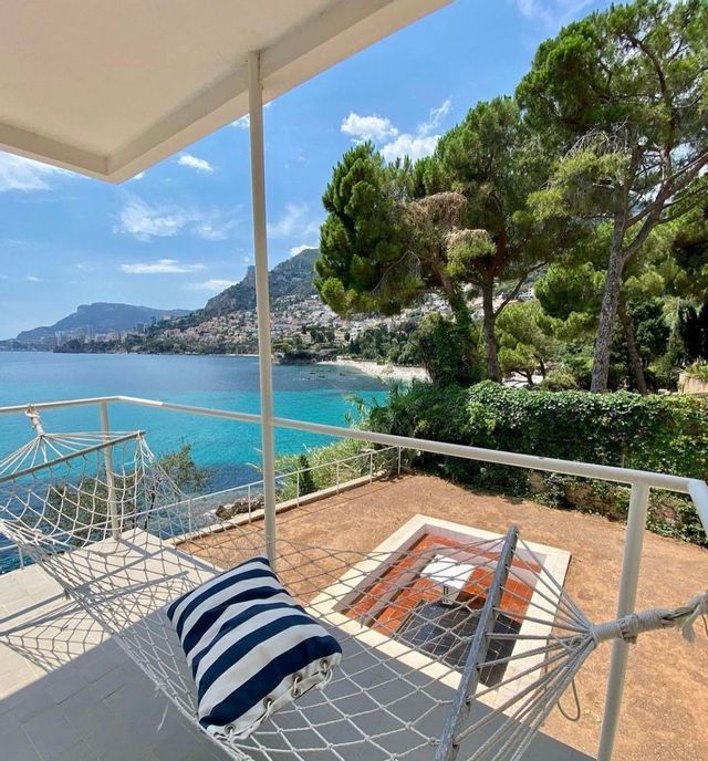 Un'amaca con un cuscino a righe bianche e blu su una terrazza con vista su una città costiera e una baia turchese.