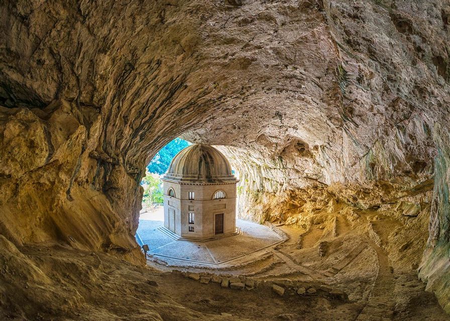 Un tempio ottagonale con cupola sorge all'interno dell'imboccatura di un'ampia grotta rocciosa, osservato dall'interno della caverna.