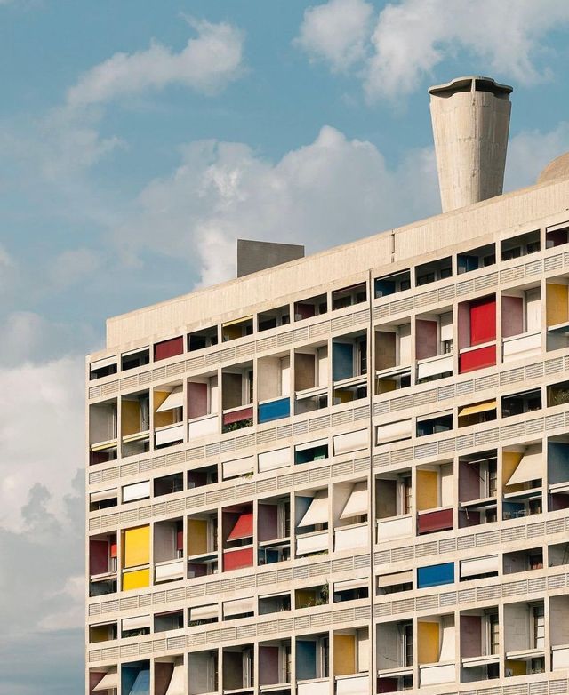 La facciata di un edificio modernista in cemento con una griglia di balconi colorati incassati, contro un cielo azzurro con nuvole.