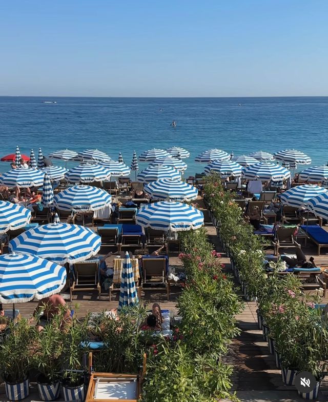 File di ombrelloni a strisce bianche e blu su una pedana di legno dove le persone si rilassano su lettini in riva al mare.
