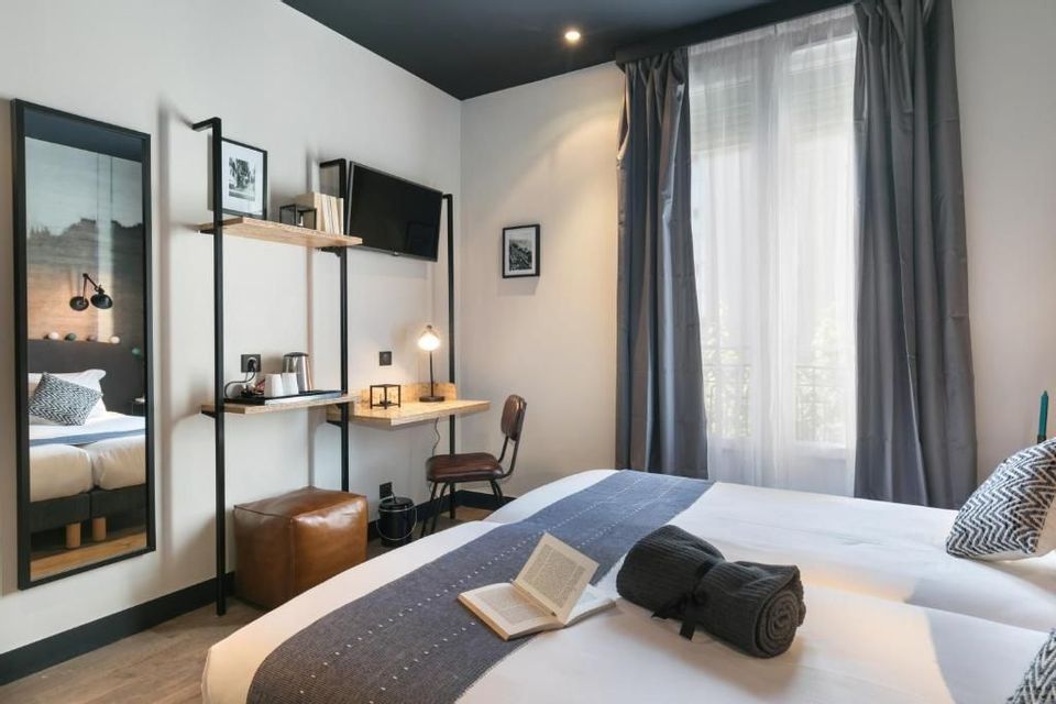 Interno di una camera d'albergo con due letti, una scrivania a muro, uno specchio e una grande finestra con tende grigie.
