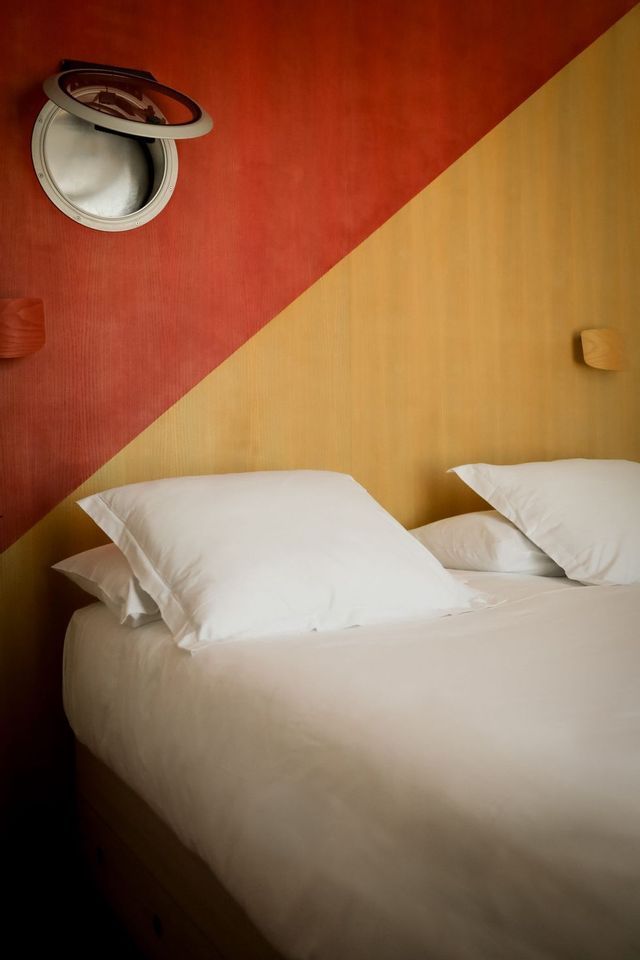 Un letto con cuscini e lenzuola bianche in una stanza con una parete dipinta in diagonale di rosso e giallo e una finestra oblò aperta.