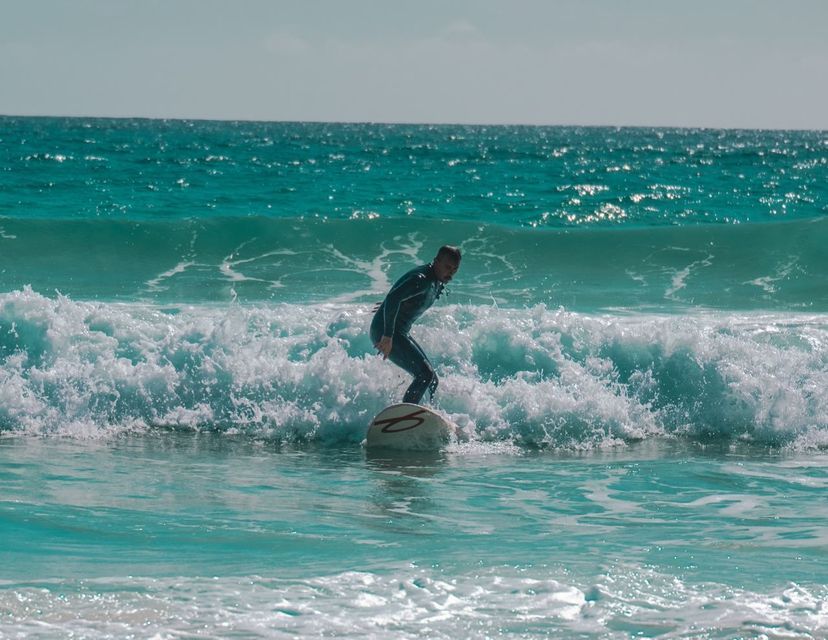 Un uomo in muta nera è in piedi su una tavola da surf, cavalcando un'onda turchese dell'oceano con schiuma bianca.
