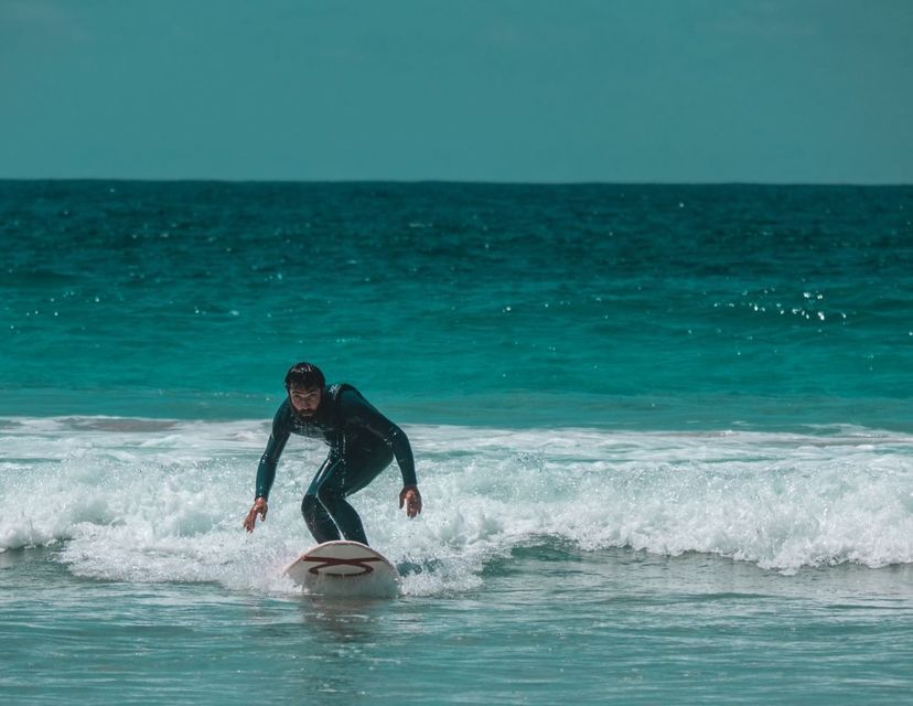 Un uomo in muta nera fa surf su una tavola bianca, cavalcando una piccola onda nel mare turchese.
