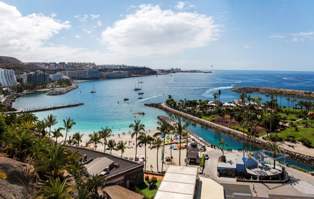 Una vista aerea della baia di una città costiera, con una spiaggia sabbiosa, palme e barche nell'acqua turchese sotto un cielo blu.