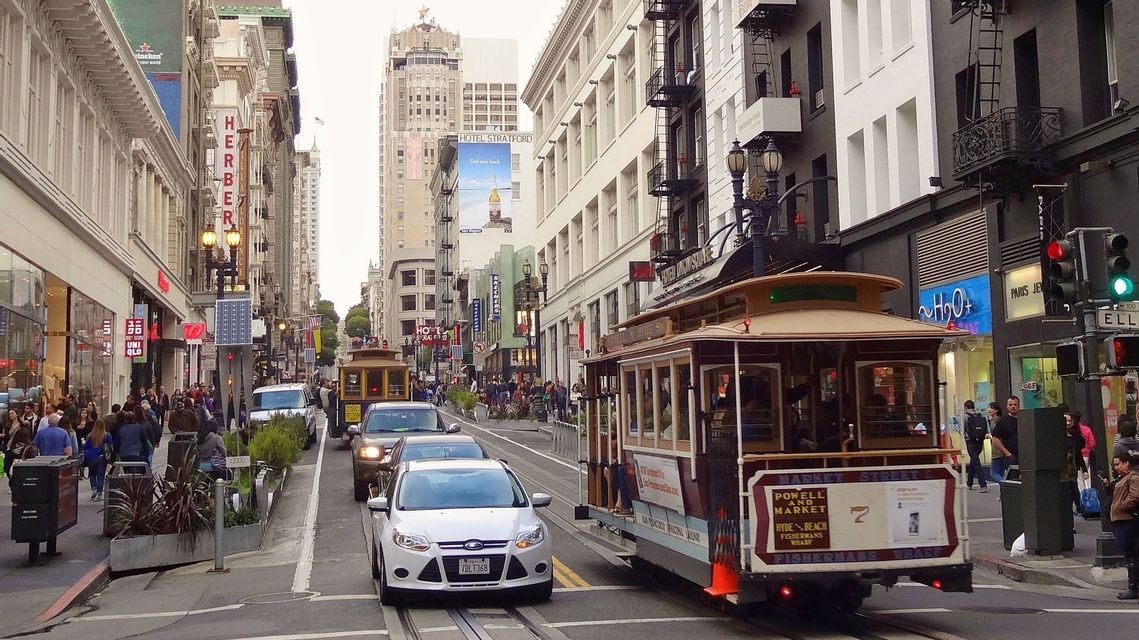 Un tranvía tradicional recorre las vías de una concurrida calle de la ciudad, rodeado de coches, peatones y edificios altos.