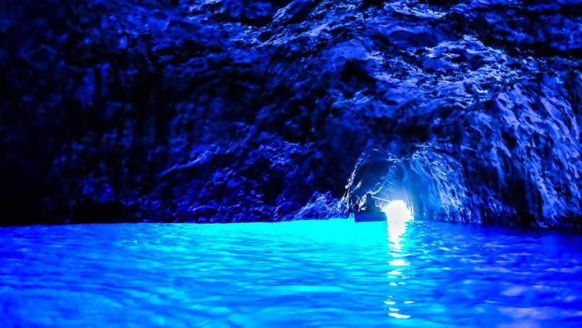 Ein kleines Boot treibt auf leuchtend blauem Wasser in einer dunklen Meereshöhle und bewegt sich auf eine sonnenbeschienene Öffnung in der Ferne zu.
