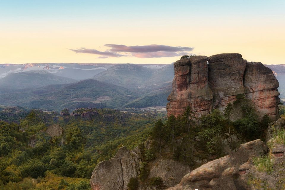 Una grande formazione rocciosa rossastra domina una valle di foreste verdi e montagne al tramonto.