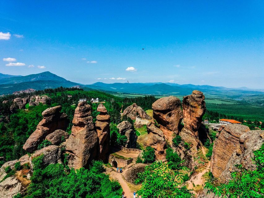 Grandi formazioni rocciose rosse si ergono da una valle verde e boscosa, con montagne distanti sotto un cielo azzurro chiaro.
