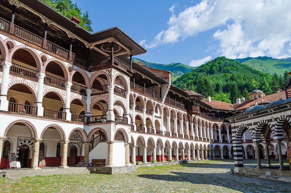 Un grande edificio con balconi ad arco su più livelli circonda un cortile acciottolato, con montagne verdi sullo sfondo.