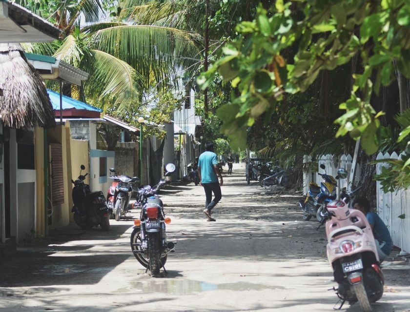 Una persona camina por una calle arenosa bordeada de motocicletas aparcadas, casas y exuberantes árboles tropicales.