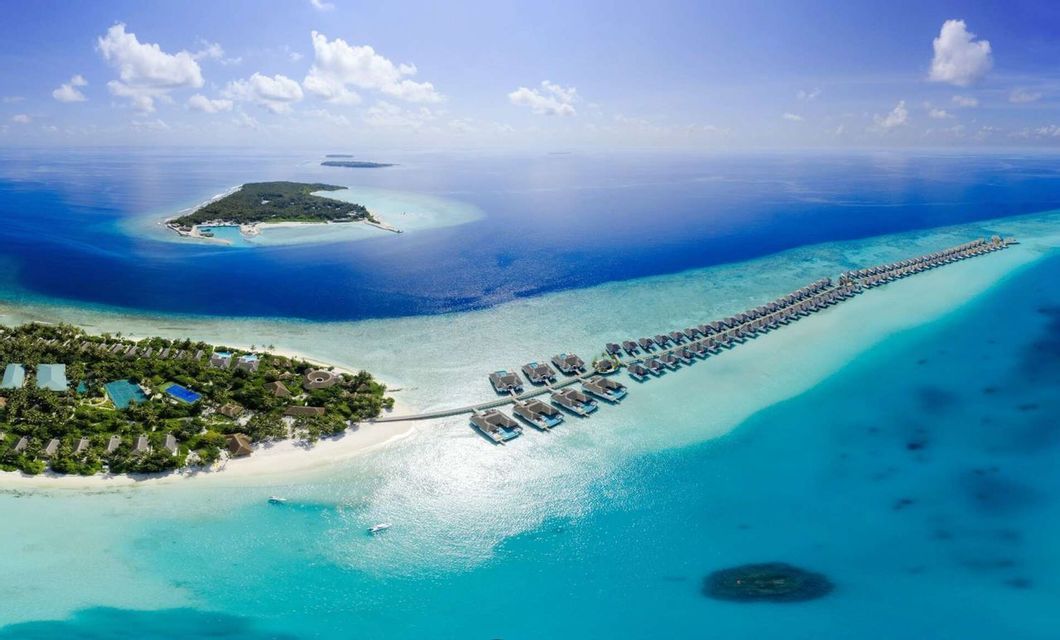 Vista aérea de un resort en una isla tropical con dos largas hileras de bungalows sobre el agua en aguas turquesas bajo un cielo azul.