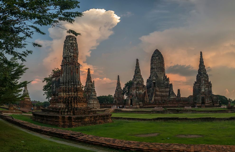 Ancient stone temple ruins with tall spires stand on a grassy field under a cloudy sky at sunset.