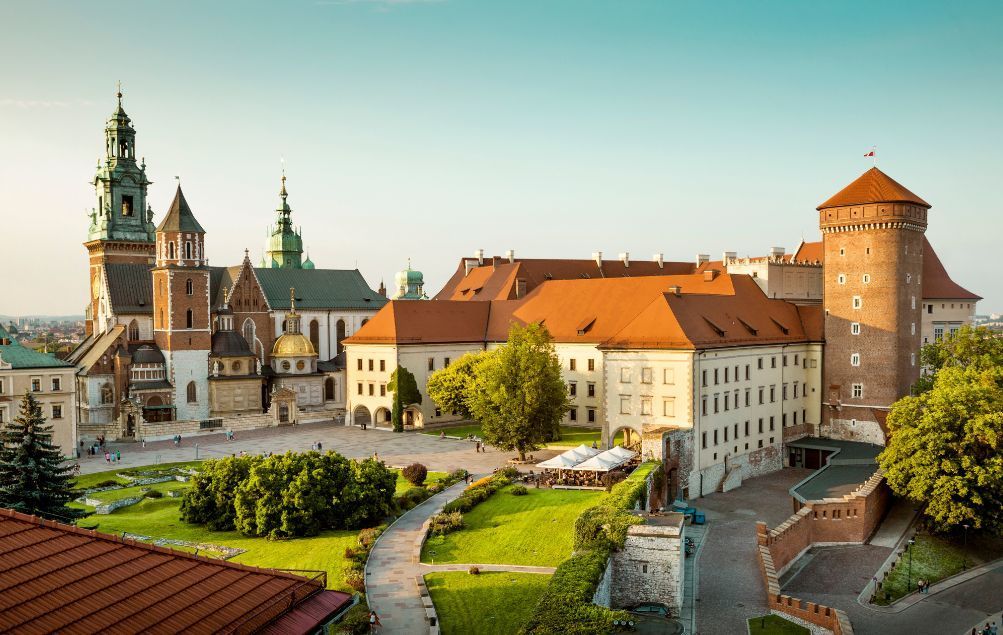 Ein erhöhter Blick auf eine große Burganlage mit einer Kathedrale, rot gedeckten Gebäuden und einem Ziegelturm, die einen Innenhof mit Gärten umschließt.