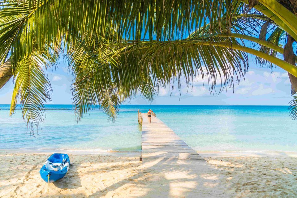 Una persona cammina su un molo di legno sopra l'acqua turchese di una spiaggia tropicale, con un kayak blu appoggiato sulla sabbia.