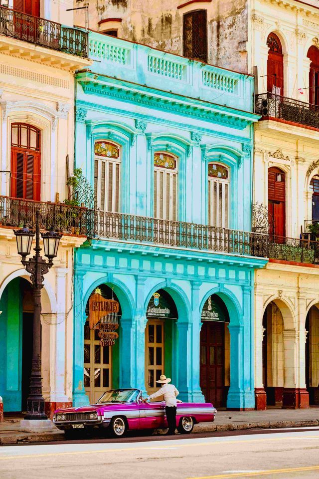 Una persona con sombrero de paja junto a un coche descapotable rosa vintage, aparcado frente a edificios coloridos y ornamentados con balcones.