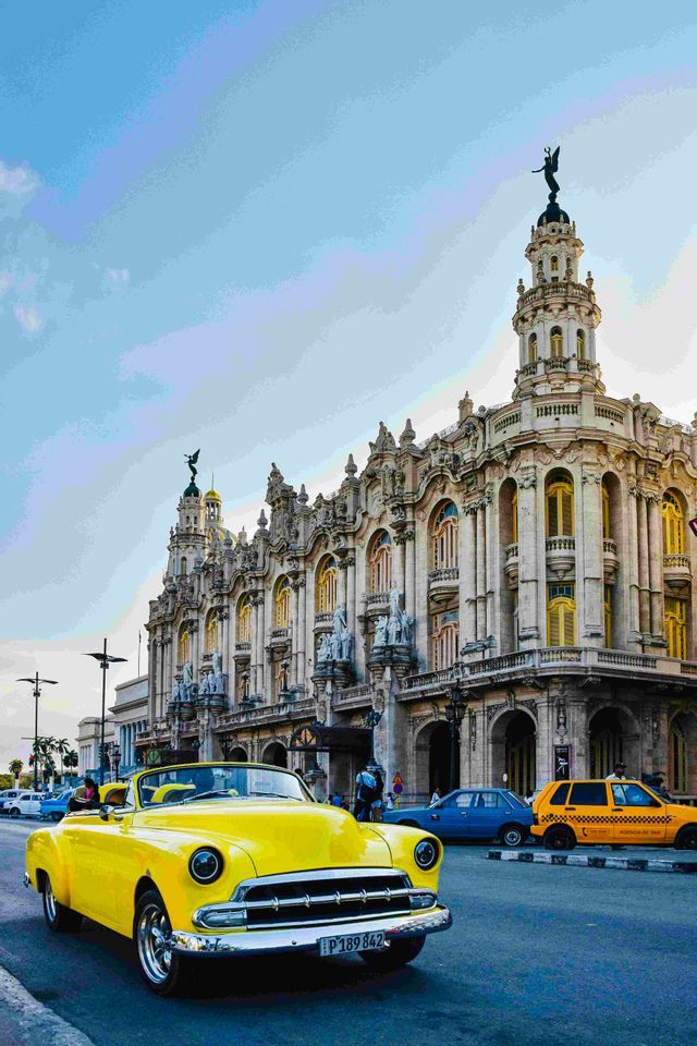Un'auto decappottabile d'epoca gialla è parcheggiata su una strada di fronte a un grande, ornato edificio storico sotto un cielo blu.