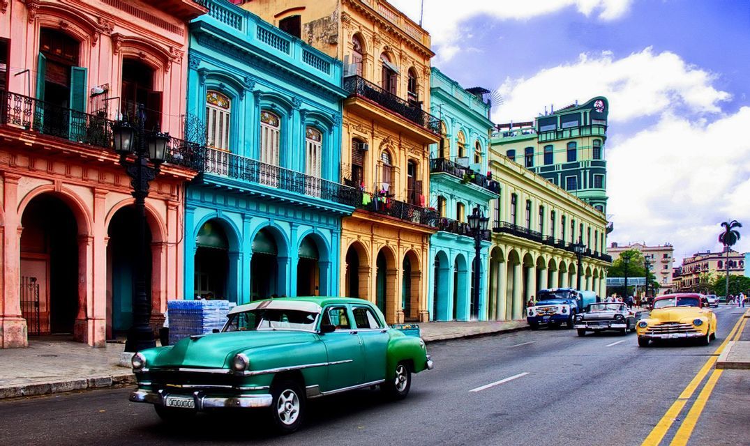 Auto d'epoca che percorrono una strada fiancheggiata da edifici coloniali dai colori vivaci con colonnati ad arco.