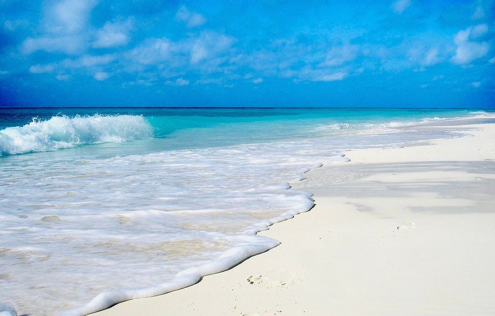 Un'onda turchese con schiuma bianca si infrange su una spiaggia di sabbia bianca sotto un cielo azzurro con nuvole soffici.