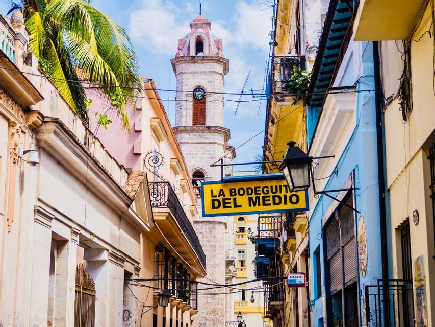 Un'insegna gialla per 'La Bodeguita del Medio' pende su una strada stretta con edifici colorati e una torre dell'orologio sullo sfondo.