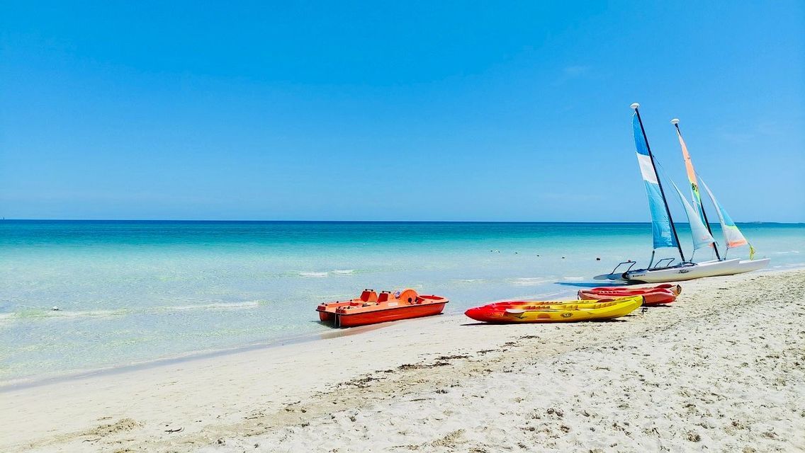 Barche a vela, kayak e un pedalò adagiati su una spiaggia di sabbia bianca accanto ad acque turchesi e limpide sotto un cielo azzurro brillante.