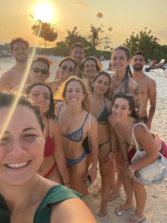 Un gruppo WeRoad in viaggio si scatta un selfie sorridente su una spiaggia sabbiosa al tramonto.