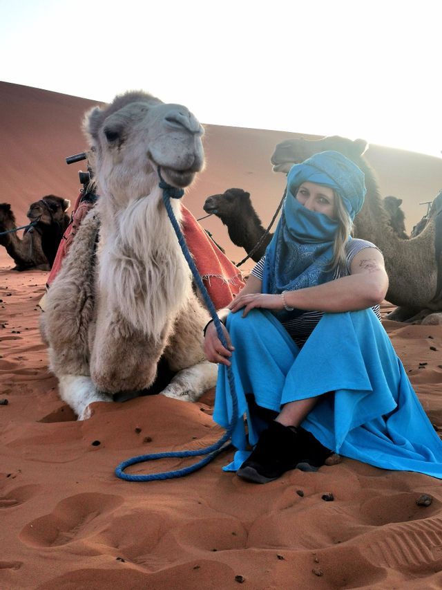 Una donna con turbante blu e vestiti siede sulla sabbia accanto a un cammello che riposa nel deserto.