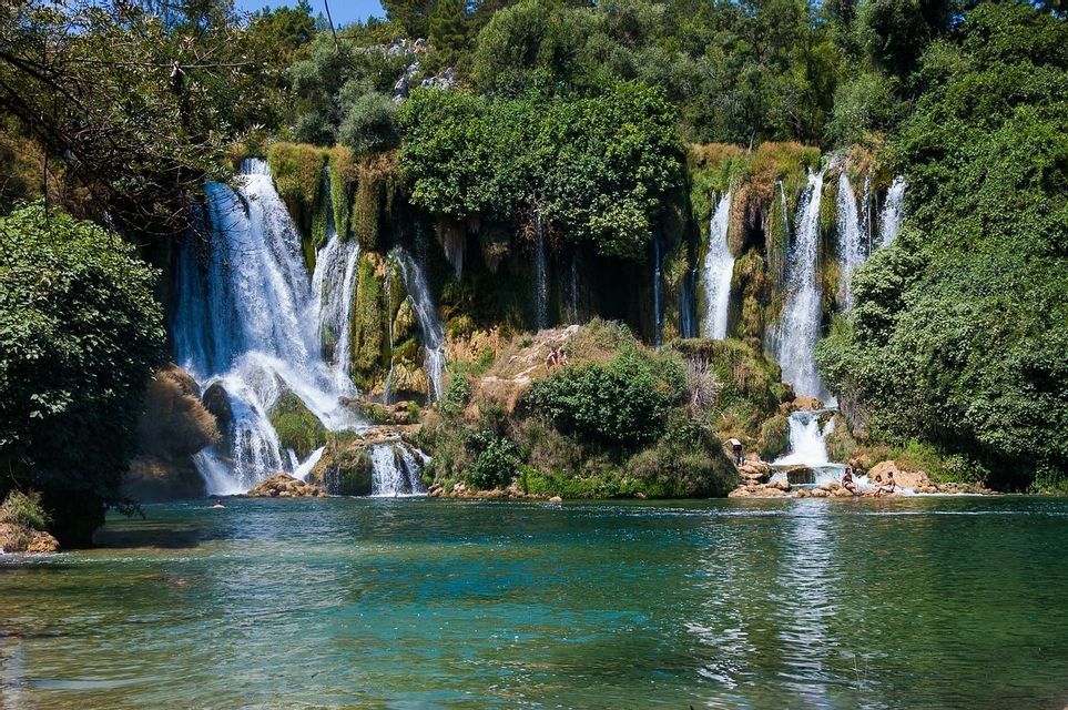 Ein mehrstufiger Wasserfall stürzt eine moosbewachsene Klippe hinab in einen türkisfarbenen Fluss, während sich Menschen auf den Felsen und im Wasser entspannen.