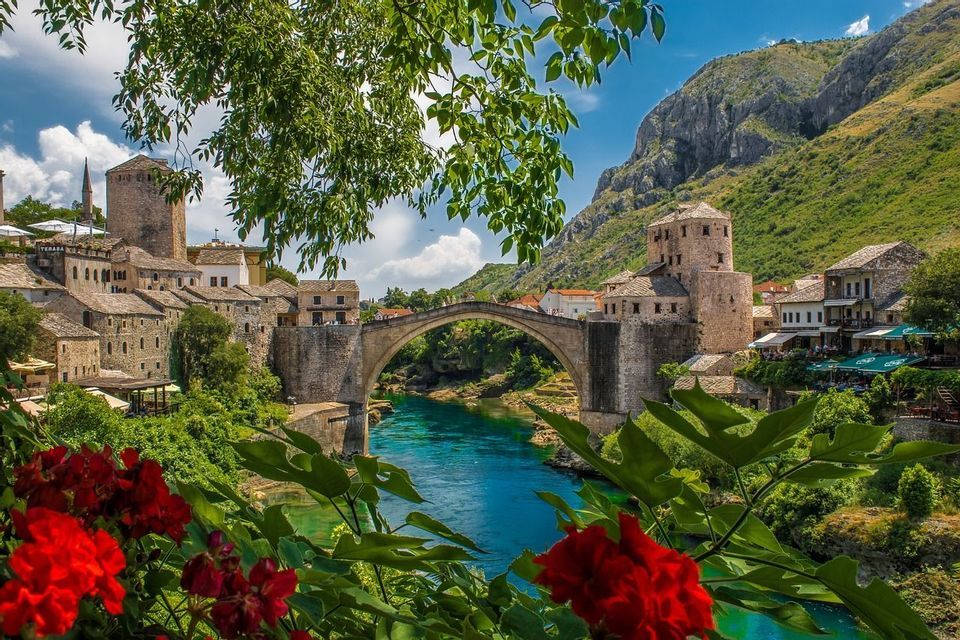 Eine historische Steinbrücke überquert einen türkisfarbenen Fluss in einer Stadt am Berghang, umrahmt von roten Blumen und grünen Blättern.