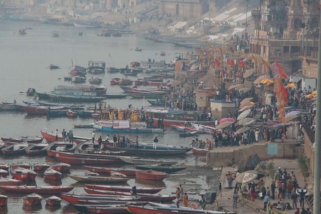 Una vista cenital de una orilla de río abarrotada, con muchas embarcaciones en el agua y personas reunidas en escalones de piedra bajo un cielo brumoso.